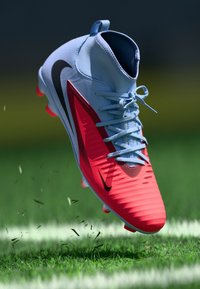 Scarpina da calcio Nike con una tomaia azzurro chiaro, accenti rossi e suola rossa testurizzata. Presenta lacci e un distintivo swoosh Nike in nero.