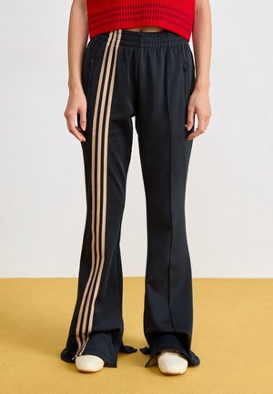 Personne portant un pantalon noir avec des bandes latérales beige et des chaussures blanches, debout sur un sol jaune sur fond blanc.