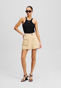 Schwarzes Tanktop mit gerippter Textur, beigefarbene Shorts mit Seitentasche und Knopfverschluss sowie schwarze Riemchen-Heels.