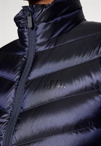 Tamsiai mėlynas pūkinis džinsas su aukštu apykakliu, horizontaliu quilted dizainu, užtrauktuku ir subtiliu juodu prekės ženklu ant krūtinės.