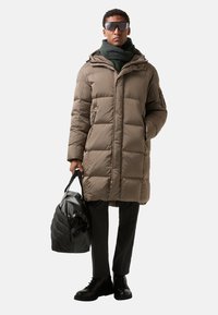 Pufferjack in taupe met een capuchon, gewatteerd patroon en rits aan de voorkant. Zwarte handtas en donkere broek, aangevuld met een groene sjaal en zonnebril.