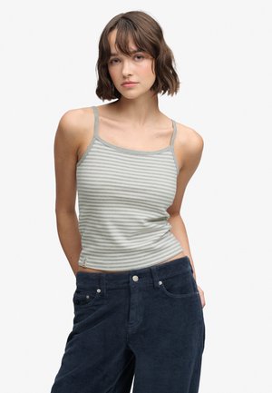 Jeune femme aux cheveux courts bruns portant un débardeur rayé gris et blanc et un pantalon en velours côtelé bleu foncé, debout les mains dans les poches.