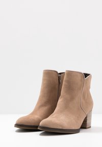 Beżowe zamszowe botki za kostkę z zaokrąglonym noskiem, bocznym zamkiem błyskawicznym i słupkowym drewnianym obcasem. Gładka tekstura i prosty design, brak widocznych wzorów.