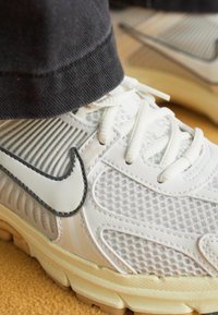 Fehér sportcipők, hálós felsőrésszel, gumitalppal és fekete részletekkel. A fűzők fehérek, a dizájnban feltűnő logó található.