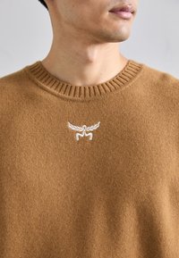 Brun strikket sweater med en ribbet rund halsudskæring. Har hvide broderede detaljer af et logo på brystet. Blød tekstur.
