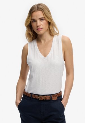 Woman with blonde hair wearing a white sleeveless V-neck top, dark pants, and a brown braided belt, hands in pockets, looking forward.