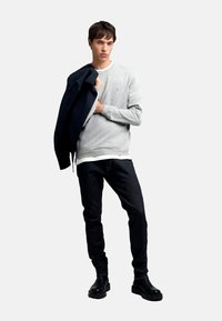 Sudadera gris claro con puños acanalados, superpuesta a una camisa blanca, combinada con jeans oscuros y botas negras hasta el tobillo. Una chaqueta azul marino está colgada sobre el hombro.