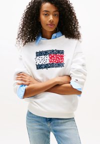 Sudadera blanca con diseño gráfico azul y rojo que presenta un patrón de bandera floral y el logotipo "TOMMY JEANS". Tejido texturizado, corte relajado.