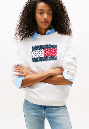 Sudadera blanca con diseño gráfico azul y rojo que presenta un patrón de bandera floral y el logotipo "TOMMY JEANS". Tejido texturizado, corte relajado.