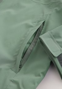 Details einer grünen wasserdichten Jacke mit einer Reißverschlusstasche und einem Bündchen mit Klettverschluss. Glatte Textur, maßgeschneidertes Design.