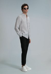 Chemise à rayures beige et brune, associée à un pantalon noir et des baskets blanches ; le modèle porte des lunettes de soleil et se tient devant un fond gris.