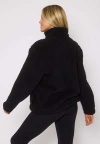 Pullover in pile nero, con colletto alto, maniche ampie e vestibilità oversize, indossato sopra leggings neri.