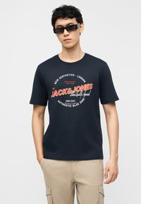 Νεαρός άνδρας που φορά μαύρα γυαλιά ηλίου και μπλε μπλουζάκι Jack & Jones με σχέδιο, φορόντας μπεζ cargo παντελόνι, στέκεται μπροστά σε απλό φόντο.