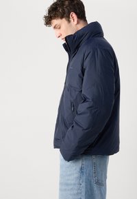 Marineblauwe gewatteerde jas met hoge kraag, rits aan de voorkant en zijzakken. Glad textuur, lichtgewicht ontwerp, geschikt voor casual kleding.