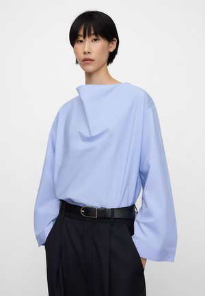 Kobieta ubrana w luźną, jasnoniebieską bluzkę z długim rękawem, włożoną do czarnych spodni z wysokim stanem i czarnym paskiem, stojąca na tle jednolitego białego tła.