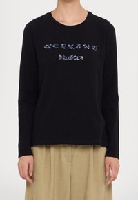 Persona che indossa una maglietta nera a maniche lunghe con il testo blu in paillettes "WEEKEND Max Mara" e pantaloni beige, in piedi contro uno sfondo neutro.