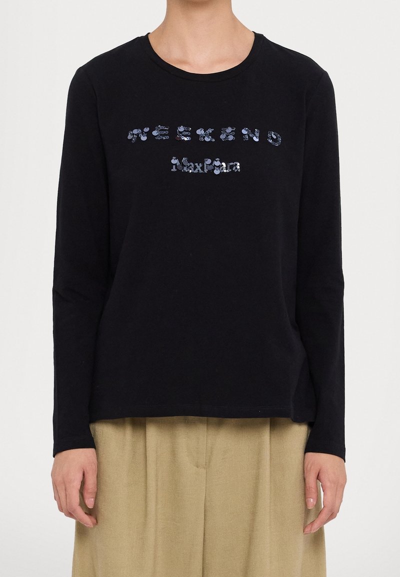 Persona che indossa una maglietta nera a maniche lunghe con il testo blu in paillettes "WEEKEND Max Mara" e pantaloni beige, in piedi contro uno sfondo neutro.