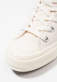Beige kanvas sneakers med en blank, hvit gummikapp og teksturert såle. Har sølvfargede snorer og hvite bomullslisser.