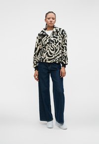 Pull en polaire dans un motif abstrait crème et noir, avec un col zippé, une poche avant et des poignets côtelés. Associé à un jean en denim à jambes larges.