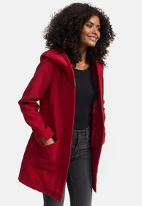 Manteau à capuche rouge en mélange de laine, avec de grandes poches, des accents de boutons marron et une coupe décontractée. Porté sur un haut noir et un jean gris.