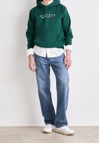 Grüne Baumwollkapuzenjacke mit Fronttasche und weißem "HILFIGER NEW YORK" Logo, übereinander getragen mit einem weißen Shirt, kombiniert mit blauen Jeans und weißen Sneakers.