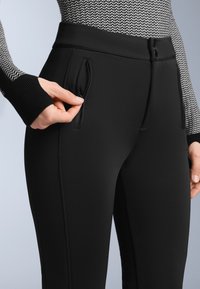 Černé sportovní kalhoty s hladkým povrchem, vybavené šikmými bočními kapsami a vysokým pasem. Šití podél švů.