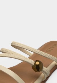 Sandale beige avec trois brides texturées, comportant une perle ronde marron brillante sur la bride du milieu, et une semelle intérieure beige avec détails de couture.