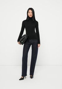 Melna turtlenecka blūze, tumši zilas platās bikses un melni smaili papēži. Modelis tur lielu melnu rokassomu, stāvot pretī baltai fona.