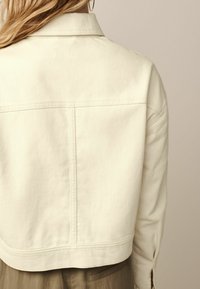 Veste en denim crème avec un design court, dotée d'un col évasé et de détails de couture visibles au dos et sur les manches.