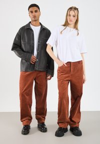 Schwarze Lederjacke mit Steppnähten, weißes T-Shirt, braune Cordhosen mit vertikalen Streifen und klobige schwarze Schuhe.