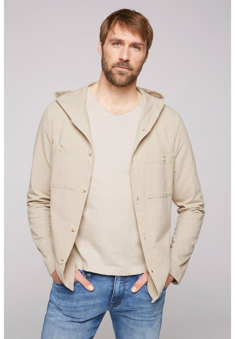 Camisa chaqueta con capucha beige, con dos bolsillos frontales, cierre de botones y mangas largas, llevada sobre una camiseta de color claro y combinada con vaqueros azules.