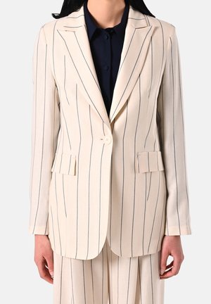 Femme portant un costume crème à fines rayures verticales noires sur une chemise noire boutonnée, debout avec les bras détendus le long du corps.