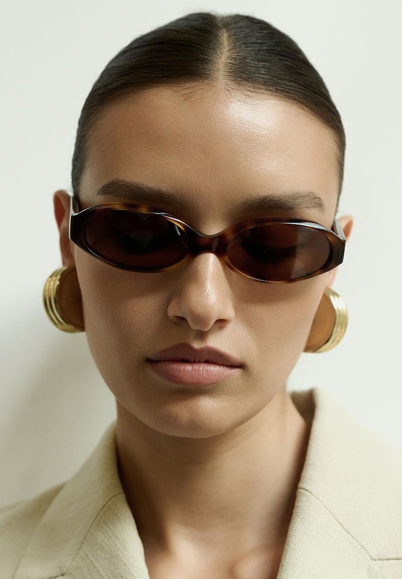 Nais, kellel on seljale kammitud juuksed, kannab kitsaid kilpkonnakuju päikseprille, ülemõõtmelisi kuldseid rõngaskõrvarõngaid ja beežikat bleiserit.