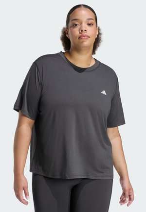 Mladá žena s kudrnatými vlasy svázanými dozadu, oblečená do tmavě šedého trička Adidas s krátkým rukávem a černých legín, stojí před světlým pozadím.