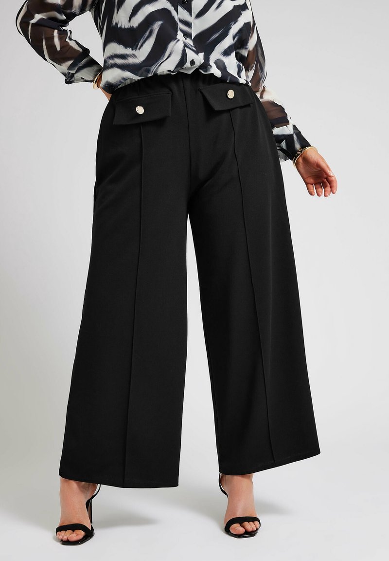 Femme portant un pantalon large noir avec des poches avant à rabat et des boutons métalliques, associé à des talons ouverts noirs et un blouse à motifs.
