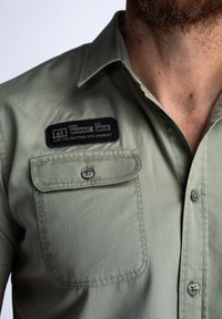Korte mouwen, olijfgroene blouse van gladde stof, met een borstzak en een zwart label met tekst en coördinaten. Twee grijze knopen.