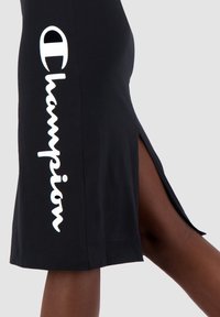 Czarna spódnica do kolan wykonana z bawełny, z bocznym rozcięciem i białym logo "Champion" w falistym wzorze po lewej stronie.