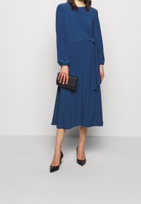 Robe portefeuille bleue en tissu doux avec des manches longues et un détail de taille nouée, assortie à une pochette noire et des escarpins pointus noirs.