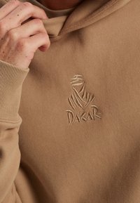 Primer plano de una mano ajustando la capucha de una sudadera beige con el logo bordado de Dakar que presenta una figura estilizada envuelta.