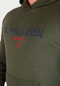 Felpa con cappuccio verde oliva realizzata in tessuto morbido. Presenta un testo ricamato in blu e rosso che recita "U.S. POLO ASSN. SINCE 1890."
