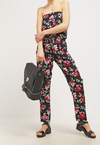 Svart blommig jumpsuit med en strapless-design, prydd med rosa och teal blommönster. Matchad med en svart handväska och klumpiga sandaler med klack.