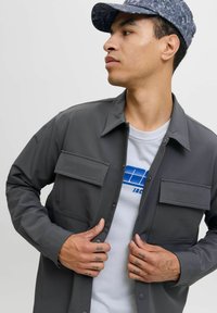 Jonge man die een donkergrijs button-up jasje over een wit T-shirt met blauwe print draagt, het jasje rechtzet terwijl hij naar de zijkant kijkt, met een gemusterde pet op.