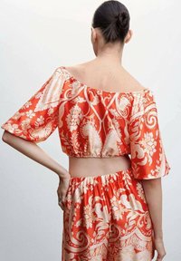 Kvinde med mørkt hår i en knold iført en rød og beige blomstrets off-shoulder crop top og matchende højtaljede bukser, vist fra ryggen.