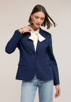 Donna con blazer blu navy sopra camicetta bianca con fiocco, jeans azzurri chiari con piccoli borchiette decorative e gioielli d'oro, che posa su sfondo semplice.