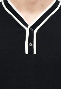 Polo de manga corta negro con cuello en V bordeado de blanco, que presenta dos botones negros y una tela texturizada.