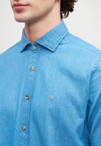 Uomo che indossa una camicia azzurro chiaro in tessuto con bottoni, con un piccolo logo ricamato sul lato sinistro del petto, in piedi di fronte a uno sfondo semplice.