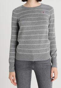 Grau gestreifter Pullover mit Rundhalsausschnitt, langen Ärmeln, geripptem Saum und einem kleinen roten Logo-Akzent auf der Brust. Aus weichem, gestricktem Material gefertigt.