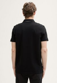 Svart polo-shirt med korta ärmar, strukturerad tyg och klassisk krage. Har sidofragor och en subtil varumärkesetikett vid fållen.