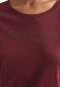 T-shirt bordeaux con colletto rotondo; presenta la stampa nera "Marc O'Polo EST. A STOCOLMA" sul petto; texture in cotone morbido.
