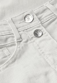 Tejido blanco a rayas con botones de metal y bordes cosidos, destacando una superficie texturizada y un detalle de bolsillo en un par de pantalones.